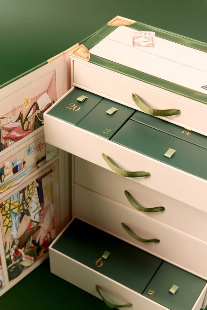 Close-up of the Vogue Festive Calendar's travel-trunk-style vanity case, showing the numbered drawers with green and cream livery and illustrated destination stickers on the lid.