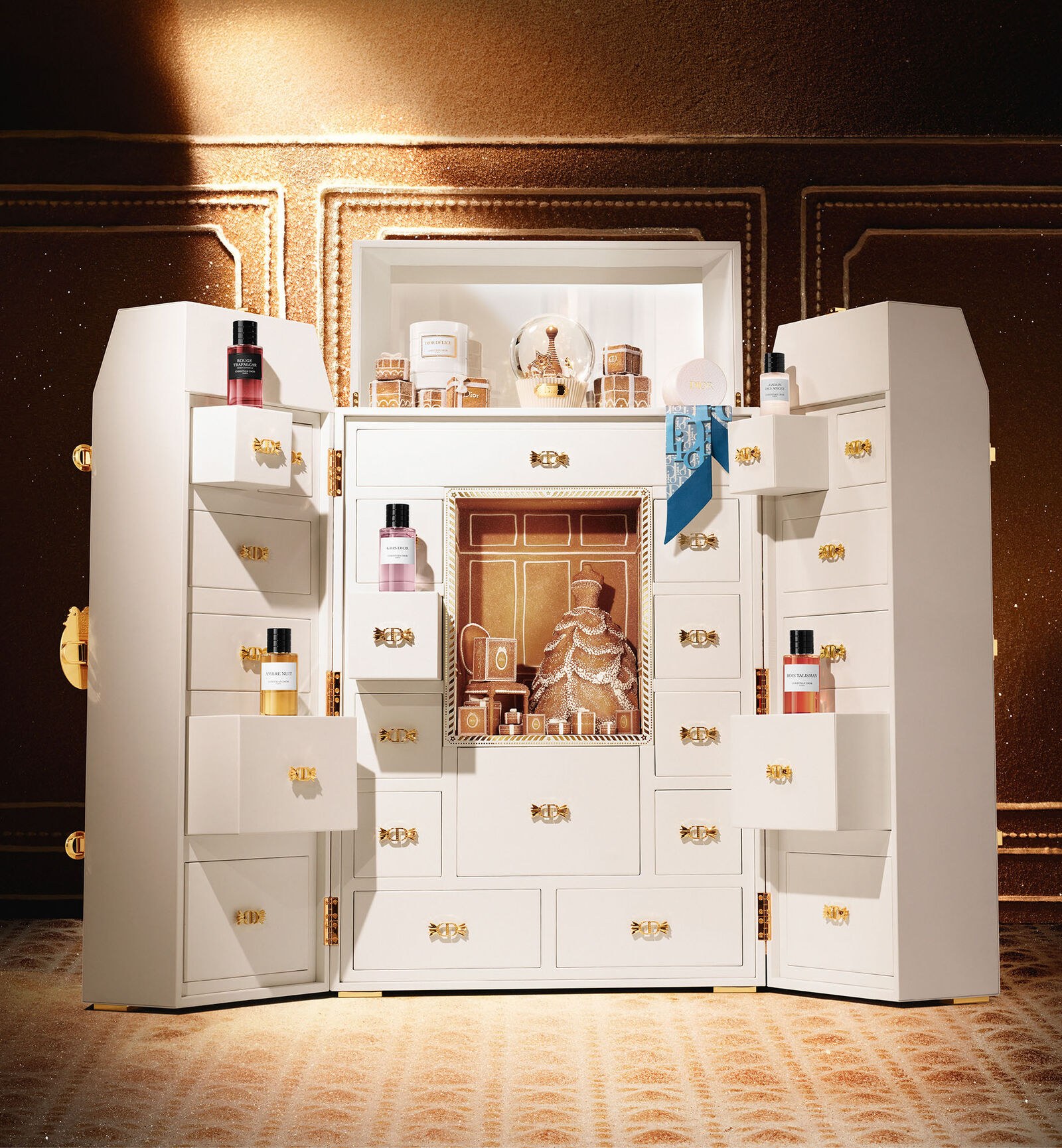The white Dior La Malle des Rêves dressing-table calendar lit against a gilded wall, open with perfume bottles and a scarf in its drawers