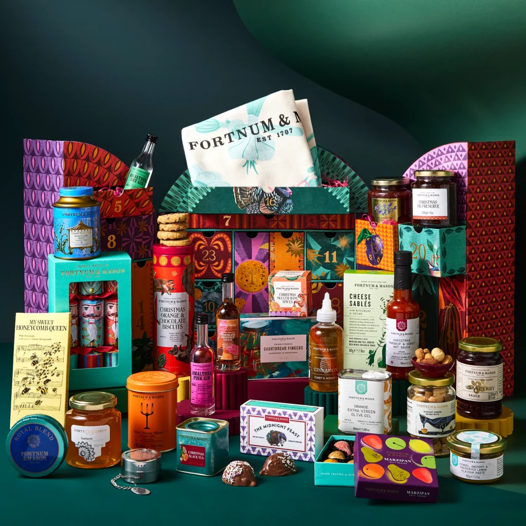 The full contents of the Fortnum’s Feasting Advent Calendar 2026 arranged in front of the open box, including preserves, tea tins, biscuits and two miniature gins.
