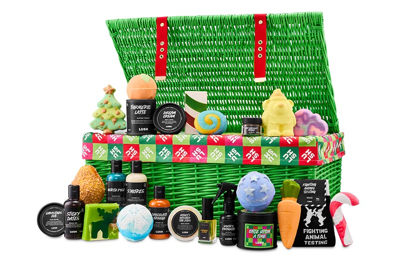 The Lush Advent Calendar 2026, an open green wicker hamper, with its bath bombs, shower gels, candle, perfume and socks arranged in front