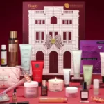 The Next Beauty Advent Calendar 2026 box with all 30 products arranged in front of it on a burgundy background