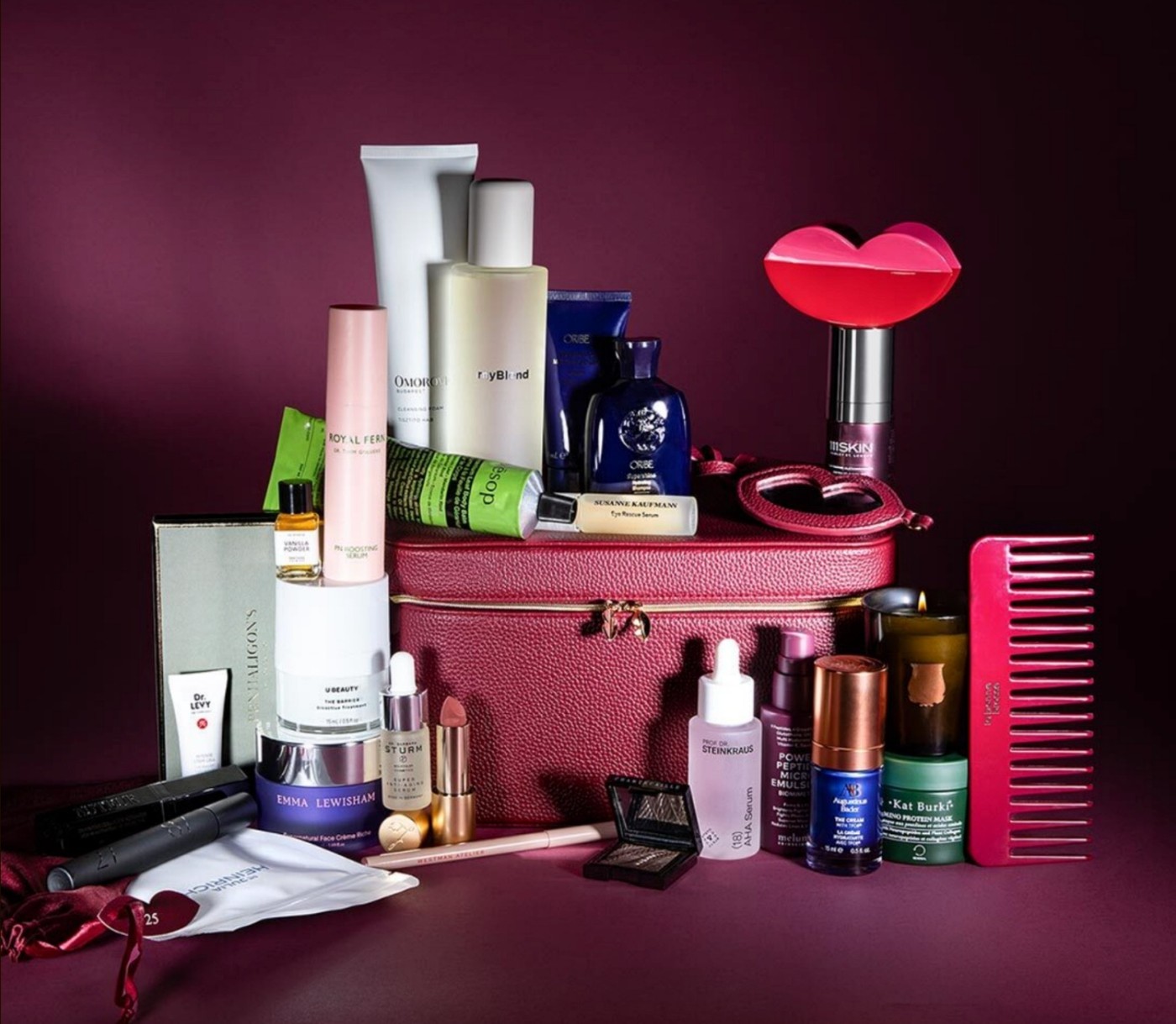 The burgundy Niche Beauty case with all the calendar products arranged around it, including a red lip-shaped hair clip, a cherry red comb, serums, creams and a candle